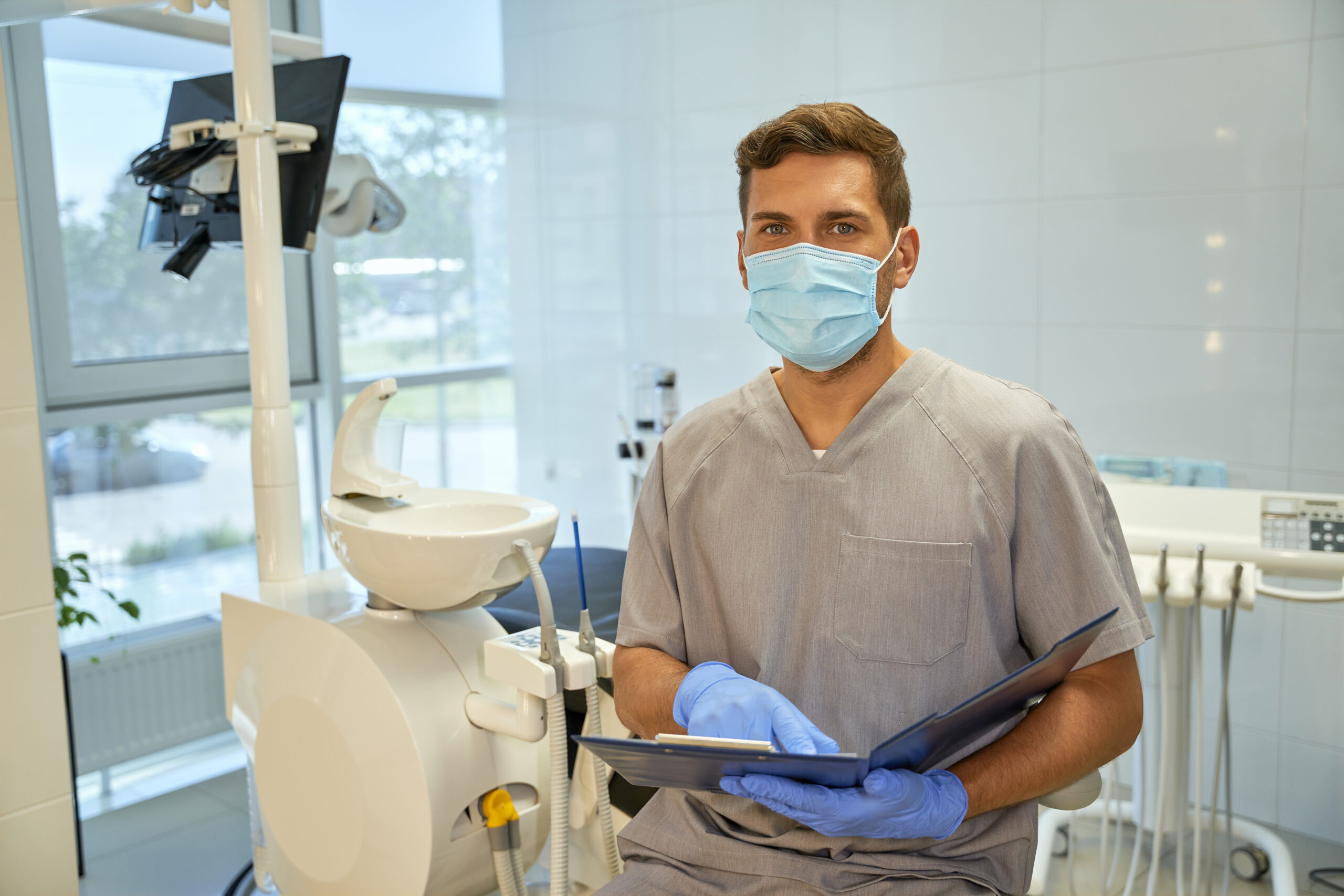 Amicable young dentist wearing a mask and holding an open folder while standing in his office