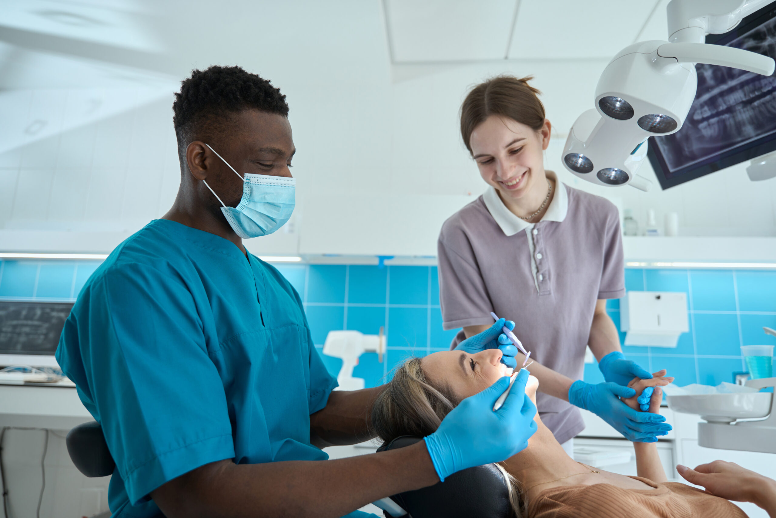 Doctor in protective mask and blue suit sitting on chair near patient and holding dental mirror